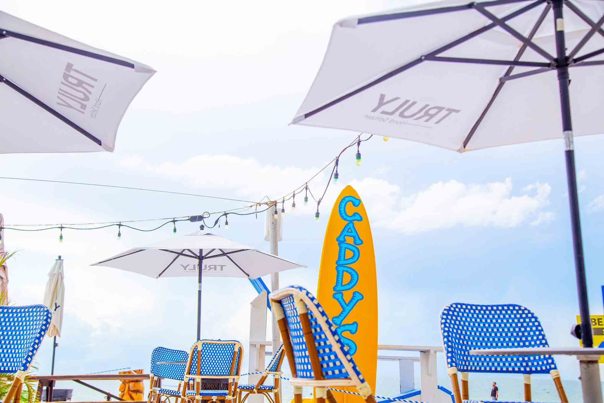 Picture of the beach at Caddy's Treasure Island with a surfboard, umbrellas, tables and blue sky